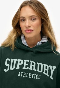 Joven mujer con cabello castaño claro que lleva puesto un hoodie de Superdry Athletics verde oscuro sobre una camisa de cuello a rayas azul y blanca.
