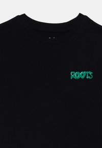 T-shirt en coton noir avec un col rond et un logo "ROOTS" vert imprimé sur le côté gauche. Texture lisse, design décontracté.