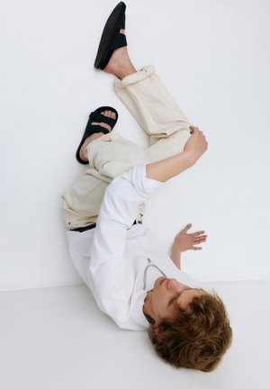 Ung person med krøllet hår iført hvid skjorte med lange ærmer, beige bukser og sorte sandaler, siddende og strækkende sig op ad en hvid væg.