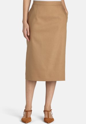 Mujer con una falda color camel de longitud hasta la rodilla con bolsillos, combinada con zapatos de tacón marrones con tira en T y punta puntiaguda, posando frente a un fondo liso.