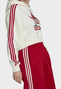 Bílá cropovaná mikina s červenými pruhy a květinovým logem, doplněná červenými kalhotami s bílými bočními pruhy. Měkká tkanina, voln ý střih.