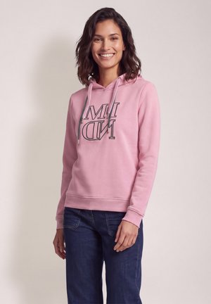 Femme souriante aux cheveux mi-longs portant un sweat à capuche rose avec un texte et un jean bleu foncé, debout devant un fond clair uni.