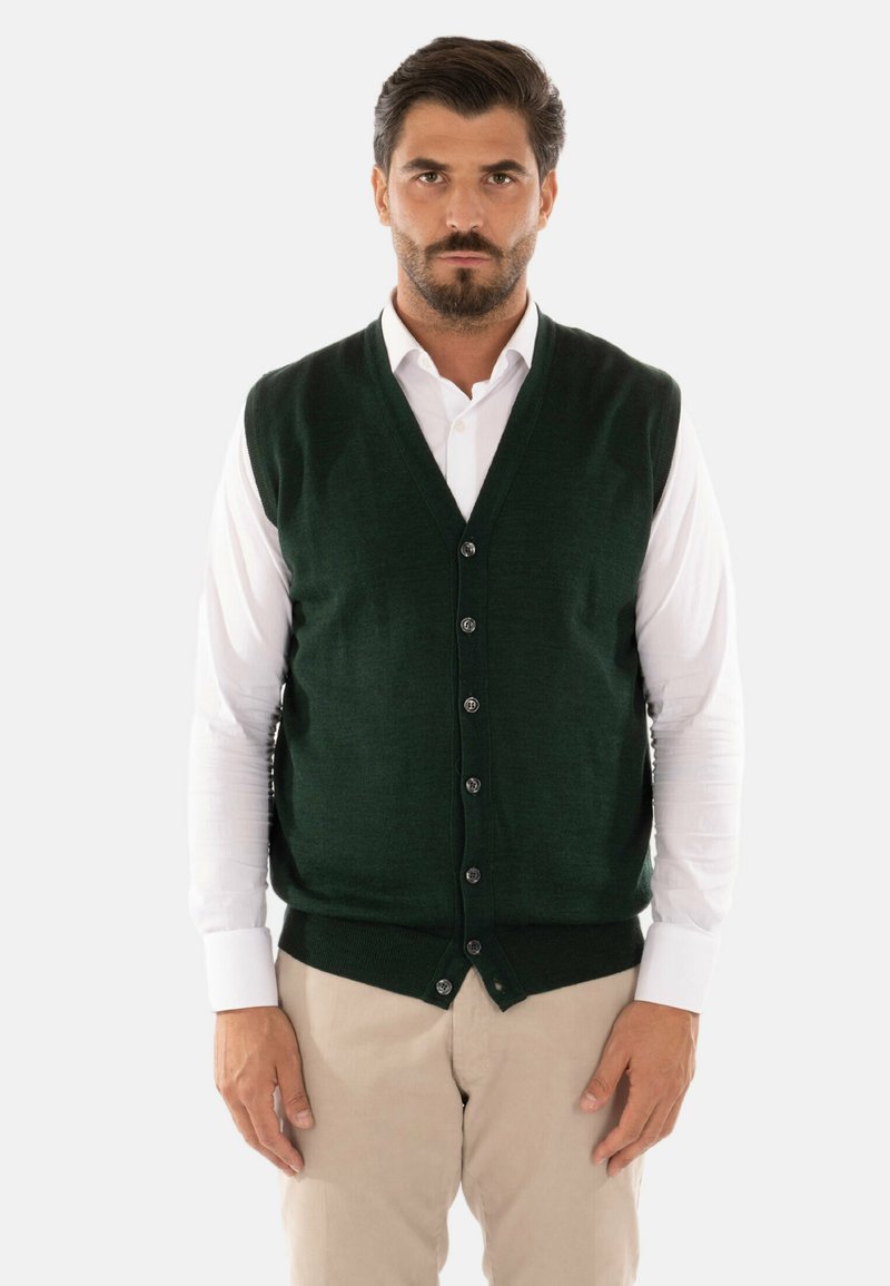 Gilet a maglia verde scuro con scollo a V, chiusura a bottoni e orlo a coste, indossato sopra una camicia bianca a maniche lunghe e pantaloni beige chiaro.