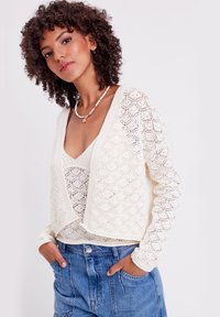 Cardigan ligero de punto crema con apertura frontal, patrón geométrico y mangas largas, llevado sobre una blusa ajustada beige. Shorts de mezclilla informales.