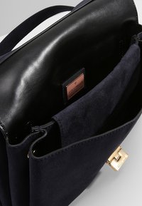 Otwarta ciemnoniebieska torebka z zamszu, z czarną skórzaną podszewką i złotym zapięciem, z widoczną metką marki "Coccinelle" w środku.