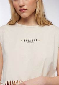 T-shirt cropped beige realizzato in morbido cotone, con scollatura rotonda, orlo a frange e testo stampato in nero "BREATHE ARIZONA" con accenti dorati.