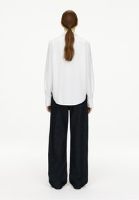 White long-sleeve shirt with a rounded hem, paired with wide dark denim trousers. The outfit features a simple, clean design.