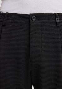 Czarne spodenki o teksturowanej powierzchni z zapięciem na guzik i szlufkami na pasek noszone na białej, włożonej koszulce.