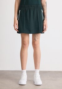 Falda atlética verde con cintura elástica, que presenta bolsillos con cremalleras blancas y un diseño de tela texturizada. Se lleva con zapatillas blancas.