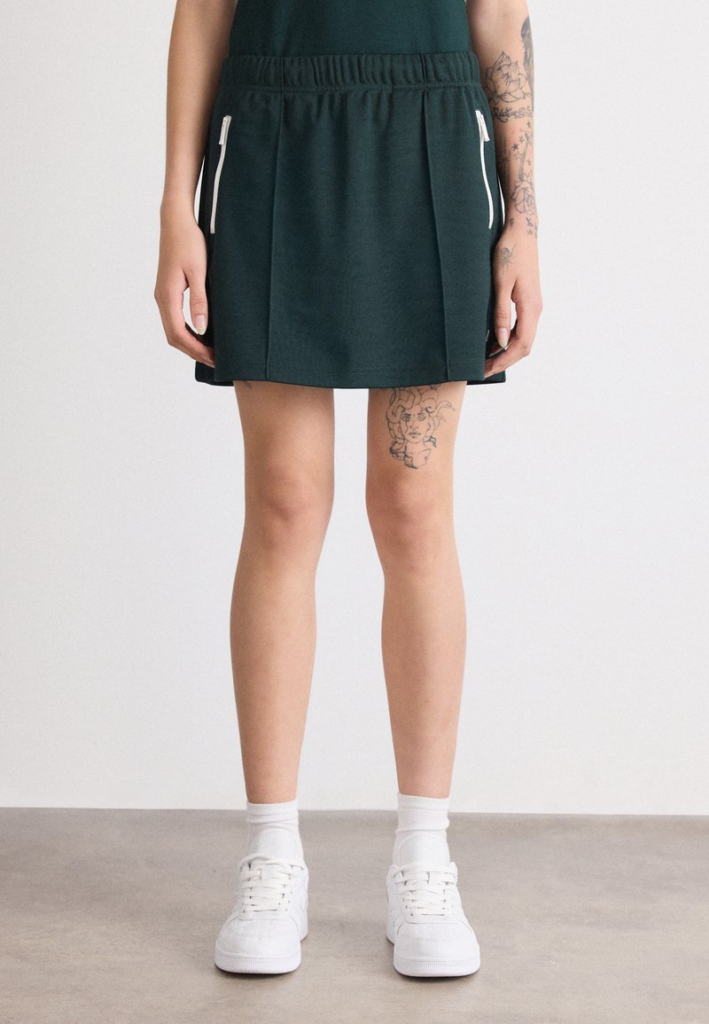 Falda atlética verde con cintura elástica, que presenta bolsillos con cremalleras blancas y un diseño de tela texturizada. Se lleva con zapatillas blancas.