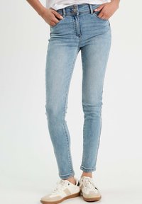 Jean skinny bleu clair taille haute, en denim, avec poches avant et fermeture éclair, associés à des baskets blanches.