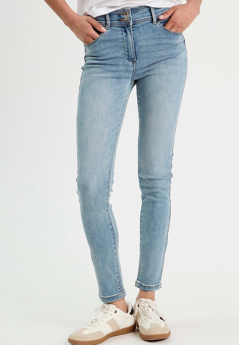 Jean skinny bleu clair taille haute, en denim, avec poches avant et fermeture éclair, associés à des baskets blanches.