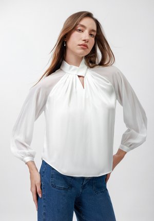 Jonge vrouw die een wit blouse met hoge hals en doorschijnende lange mouwen draagt, gecombineerd met een blauwe spijkerbroek, poseert met een hand op haar heup tegen een effen achtergrond.