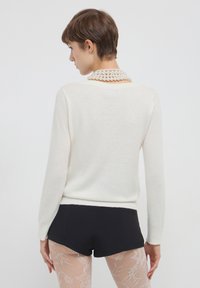 Donna con capelli corti castani che indossa un maglione bianco a maniche lunghe, pantaloni corti neri, calze a rete e una collana di perle a più fili, di spalle.