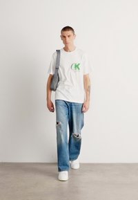 Camiseta blanca con logo verde, jeans azules desgastados y zapatillas blancas. Bolsa cruzada gris colgada del hombro. Ambiente minimalista.
