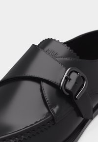 Chaussure habillée en cuir noir avec une bride de moine et une boucle argentée, présentant une finition lisse et polie en gros plan.