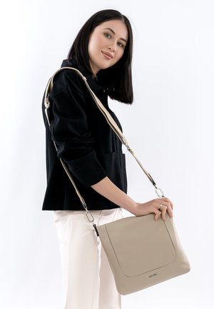 Junge Frau trägt schwarze Jacke und weiße Hose und hält eine beige Umhängetasche aus Leder mit verstellbarem Riemen.