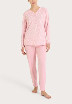 Donna che indossa una maglia a maniche lunghe rosa tenue con motivi floreali e bottoni, abbinata a pantaloni rosa tinta unita, in piedi a piedi nudi su uno sfondo chiaro.