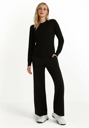 Suéter de tejido negro de manga larga combinado con pantalones anchos negros. El modelo lleva zapatillas blancas, mostrando una combinación de atuendo casual.