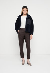 Chaqueta de felpa azul marino con un bolsillo frontal, parte superior blanca y pantalones marrones ajustados. La modelo lleva zapatos planos beige con punta, de pie contra un fondo neutro.