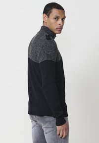 Suéter de punto negro y gris con textura acanalada, cuello con medio zipper y detalles de botones en los hombros. Corte ajustado con diseño de bloques de color contrastantes.