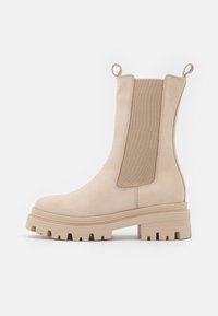 Beige suede ankle boot with elastic side panels, round toe, and chunky rubber sole featuring tread patterns. Integrated pull tabs for easy wear.