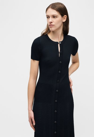 Γυναίκα με μακριά καστανά μαλλιά, που φοράει ένα μαύρο πλεκτό φόρεμα με κοντά μανίκια και κουμπιά, με υφασμάτινα σχέδια, στέκεται μπροστά σε ένα απλό φωτεινό φόντο.