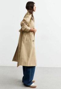 Beige trenchcoat met knopen, zijsplitten en brede mouwen, gedragen over blauwe wijde jeans en crèmekleurige sneakers op een lichtgrijze vloer.