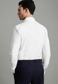 Cămașă albă cu nasturi, mâneci lungi, croi slim, asociată cu pantaloni în nuanța bleumarin închis. Material neted, linii clare și detalii subtile de cusături.