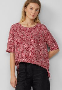 Rood gemusterde blouse met korte mouwen, een ronde halslijn en zijkanten met strikjes; heeft diamanten vormen in het wit tegen een rode achtergrond.