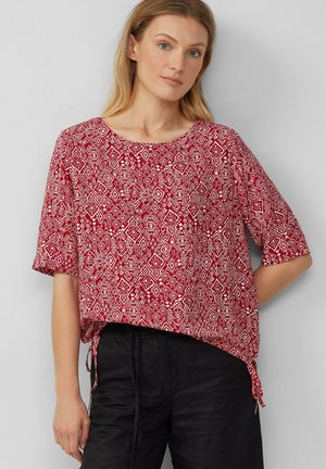 Rot gemusterte Bluse mit kurzen Ärmeln, einem runden Ausschnitt und seitlichen Bindebändern; zeigt diamantartige Formen in Weiß auf rotem Hintergrund.