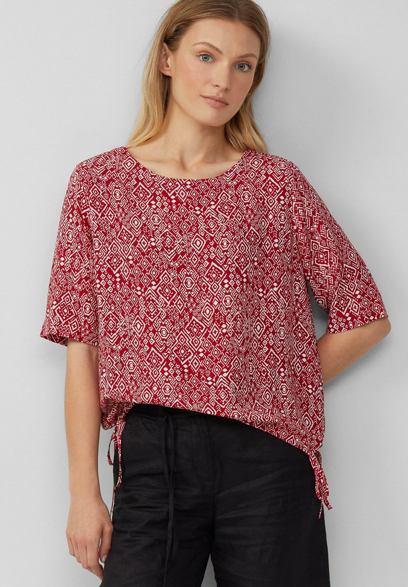 Rood gemusterde blouse met korte mouwen, een ronde halslijn en zijkanten met strikjes; heeft diamanten vormen in het wit tegen een rode achtergrond.