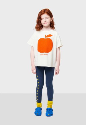 Weißes T-Shirt mit orangefarbigem Apfelmotiv, blaue Leggings mit gelbem Text, gelbe Socken und blaue Clogs. Lässiges Outfit für Kinder.