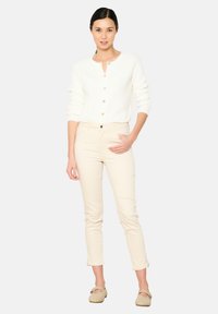 Bílý žebrovaný cardigan s knoflíkovou légou, spárovaný se světle béžovými slim kalhotami a krémovými slip-on botami. Celkově jednoduchý design.