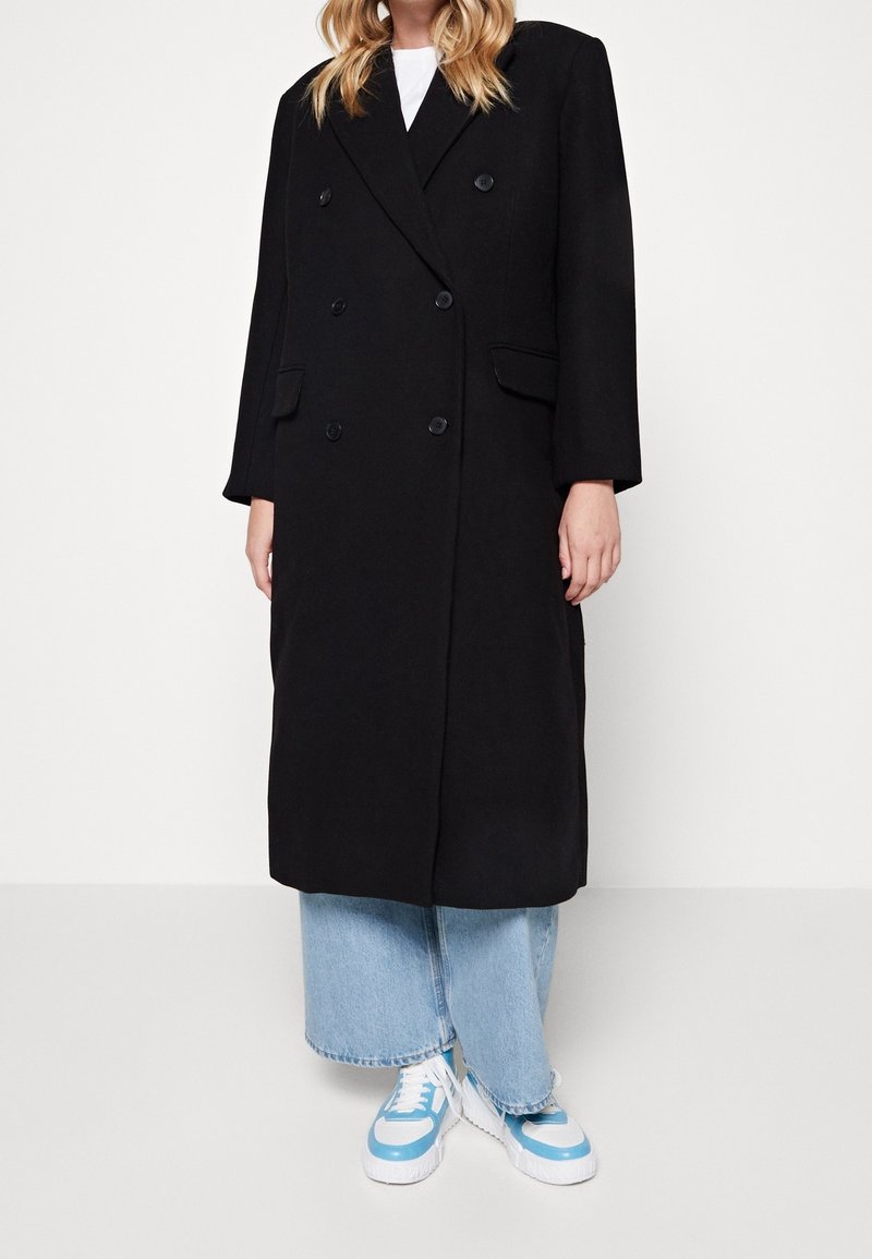 Abrigo negro de doble pecho con cuello con muescas, dos bolsillos frontales y ajuste holgado. Combinado con jeans de mezclilla azul claro y zapatillas blancas.