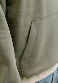 Oliivinvihreä neulospaita pehmeällä tekstuurilla, jossa on etutasku ja resoreilla viimeistelty helma. Valkoisen paidan päällä, kuvattu sivulta päin.