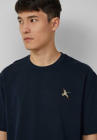 Junger Mann in einem navyblauen T-Shirt mit einem kleinen gestickten fliegenden Entenmotiv auf der linken Brust, der nach links schaut, vor einem schlichten Hintergrund.