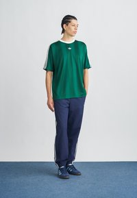 Πράσινο κοντομάνικο πουκάμισο με λευκό γιακά, τρεις λευκές ρίγες κατά μήκος των μανικιών. Συνδυασμένο με μπλε-σκούρο παντελόνι και μπλε αθλητικά παπούτσια.