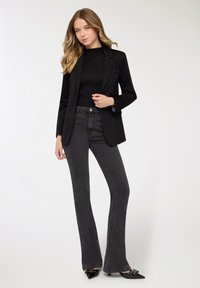 Blazer nero con una vestibilità su misura, abbinato a jeans svasati grigio scuro. L'outfit presenta una texture liscia e décolleté nere con punta.