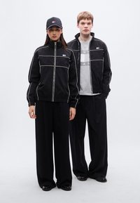 Μαύρα μπουφάν με φερμουάρ και λευκές γραμμές, συνδυασμένα με άνετα μαύρα παντελόνια και μαύρο καπέλο. Απλός σχεδιασμός και άνετη εφαρμογή.