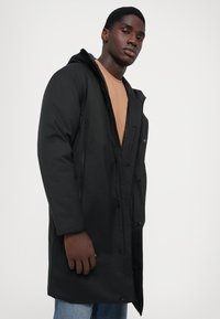 Parka nera con cappuccio, dotata di zip e chiusura a bottone, tasche frontali e vestibilità rilassata. Realizzata in un tessuto liscio e opaco.