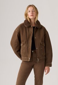 Jonge vrouw in een bruine spijkerjasje met een shearling kraag en bijpassende bruine broek, staand voor een eenvoudige lichte achtergrond.