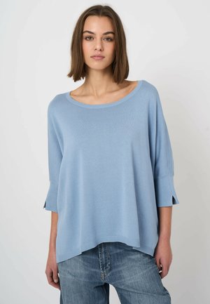 Kobieta o prostych brązowych włosach ubrana w luźny, jasnoniebieski sweter z rękawami do łokcia oraz niebieskie dżinsy, stoi na jednolitym tle.