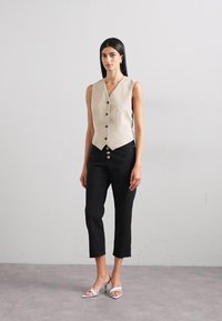 Gilet in lino beige con scollo a V e cinque bottoni neri, abbinato a pantaloni neri a vita alta e sandali bianchi con tacco. Design semplice e aderente.