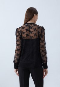 Black lace blouse with sheer fabric, floral pattern, long sleeves, and a buttoned back opening. Features a solid black underlayer.