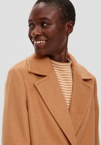 Camelkleurige wolblendjas met een notch-kraag. De stof is zacht, met een gladde textuur en een licht oversized pasvorm, gedragen over een gestreepte top.