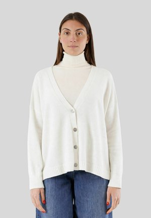 Donna che indossa un cardigan bianco abbottonato sopra un dolcevita panna e jeans blu, in piedi davanti a uno sfondo grigio uniforme.
