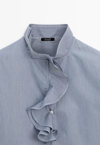 Modrá a bílá tence pruhovaná košile s vysokým límečkem a řasenou přední légou, viditelný jeden malý bílý knoflík.