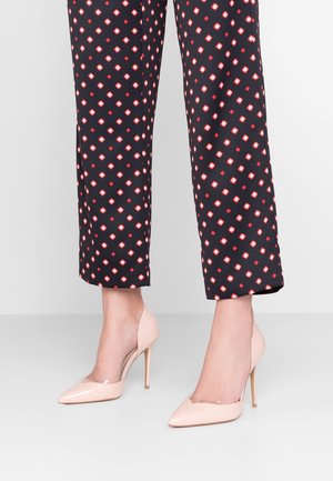 Beżowe szpilki na spiczastym nosku o błyszczącej powierzchni z przezroczanym bokiem, w zestawieniu z wzorzystymi spodniami w lina o czerwono-białych rombach.