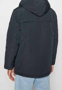 Marineblauwe gewatteerde jas met capuchon, voorzien van een gladde textuur en een detail op de achterkant. Lange mouwen en een licht gebogen zoom.